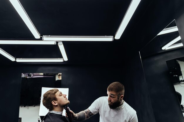Peluquero tatuado recortando hombre barbudo con máquina de afeitar en barbería Proceso de peluquería Peluquero cortando la barba de un hombre barbudo