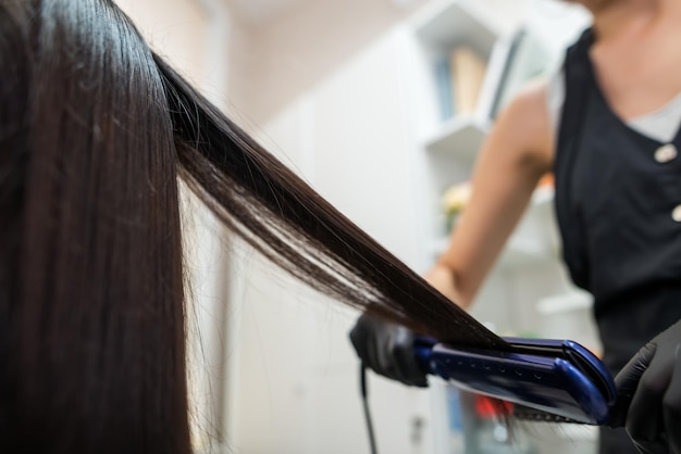 Peluquero usando un cabello alisado para alisar el cabello Estilista trabajando en el peinado de una mujer en el salónCuidado del cabello Hermosa mujer atractiva en un salón de belleza