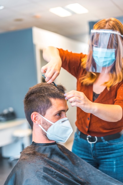 Peluqueros después de la pandemia de coronavirus. Peluquería caucásica con mascarilla y pantalla protectora, covid-19. Distancia social, nueva normalidad. Cortar el cabello con unas tijeras a una joven morena