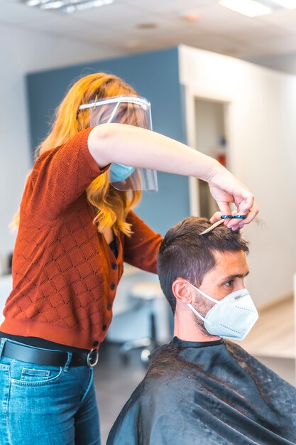 Peluqueros después de la pandemia de coronavirus. Peluquería con mascarilla y pantalla protectora, covid-19. Distancia social, nueva normalidad. Cortar el flequillo de un joven con unas tijeras, foto vertical