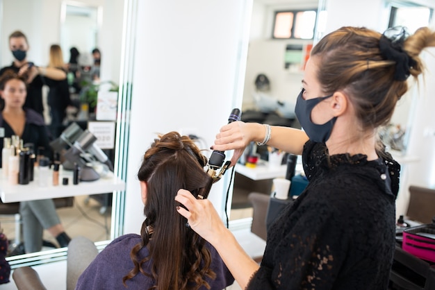 Peluqueros enmascarados que trabajan en el cabello y el maquillaje de un cliente, trabajan durante el coronavirus y el concepto de moda.