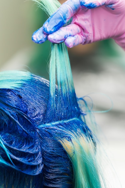 Los peluqueros con la mano en el guante protector levantan el choque del cabello azul moderno del cliente durante el proceso de teñido del cabello en un salón de belleza profesional.