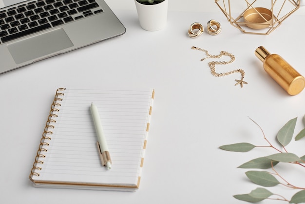 Pendientes de oro y cadena, cuaderno con bolígrafo, botella de aroma y teclado portátil en el escritorio blanco que es el lugar de trabajo del empleado