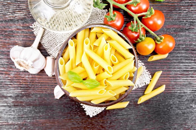 Penne-Nudeln aus Weizenmehl in einer Schüssel mit Kokosnussschalen auf Tomaten, Knoblauch-Pflanzenöl in einem Dekanter und Petersilie auf Holzbretthintergrund von oben