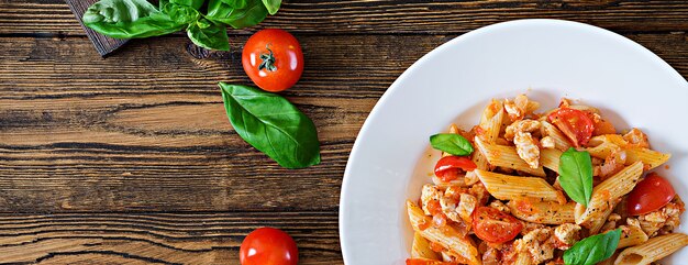 Penne pasta en salsa de tomate con pollo, tomates, decorada con albahaca sobre una mesa de madera. Comida italiana. Pasta boloñesa. Vista superior. Lay Flat