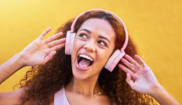 Pensamiento musical y asombro con una mujer negra escuchando la radio al aire libre en un fondo de pared amarilla Energía de auriculares y cara con una atractiva joven transmitiendo sonido de audio para divertirse