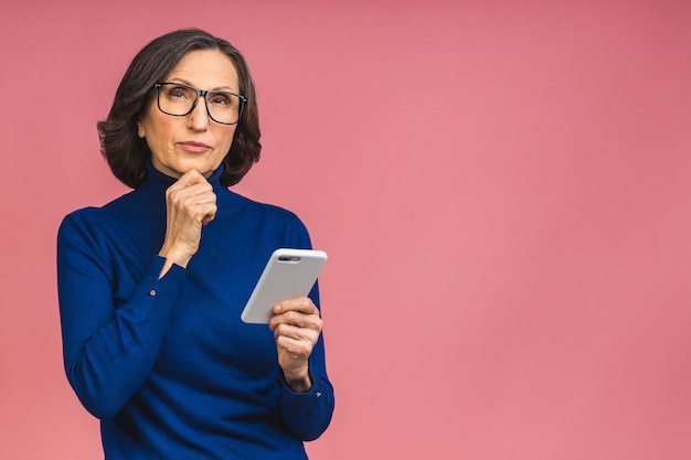 Pensamiento serio mujer senior madura sosteniendo smartphone usando aplicaciones móviles en línea, sonriendo anciana de mediana edad enviando mensajes de texto sms charlando en el teléfono aislado sobre fondo rosa.