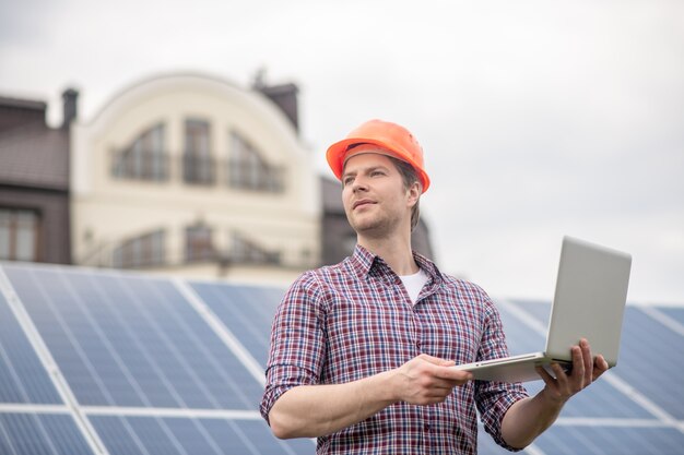 Pensamientos, idea. Hombre en casco protector con portátil abierto mirando pensativamente al lado de pie contra el fondo de la batería solar cerca de la nueva casa