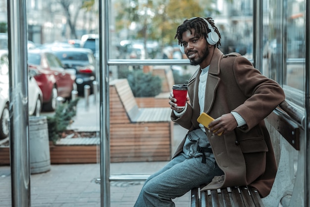 Pensamientos positivos. Encantado de hombre barbudo sosteniendo un vaso de papel y buscando autobús