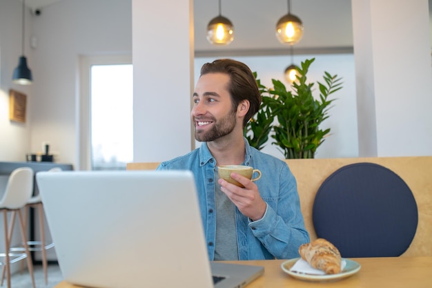 Pensamientos positivos. Hombre adulto joven feliz con café sentado en la mesa con una computadora portátil felizmente mirando hacia otro lado soñadoramente