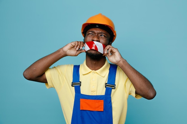 Pensando a boca selada com fita jovem construtor afro-americano de uniforme isolado em fundo azul