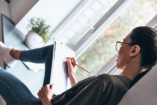 Pensando en cada detalle. Vista superior de la mujer joven concentrada escribiendo algo mientras está sentado en el alféizar de la ventana en la oficina