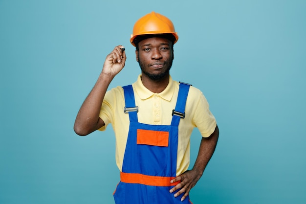 pensando colocando a mão nos quadris jovem construtor americano africano em uniforme segurando marcador isolado em fundo azul