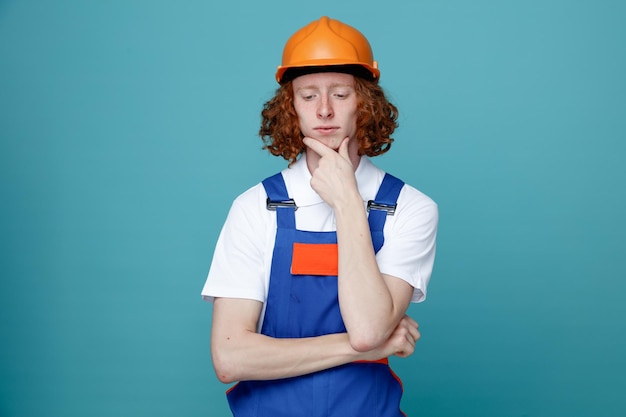 Pensando com os olhos fechados agarrou o queixo jovem construtor de uniforme isolado em fundo azul