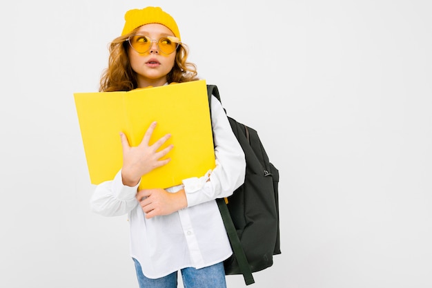 Pensando elegante caucasiano encantadora colegial em uma camisa branca, com um chapéu e uma mochila com um caderno nas mãos em branco