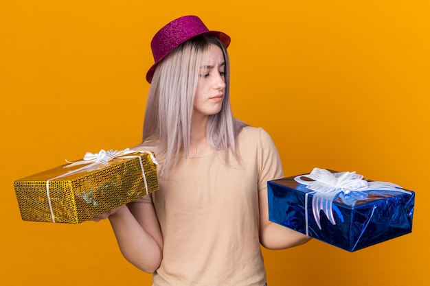 Pensando em uma jovem mulher bonita com chapéu de festa segurando e olhando para caixas de presente isoladas na parede laranja