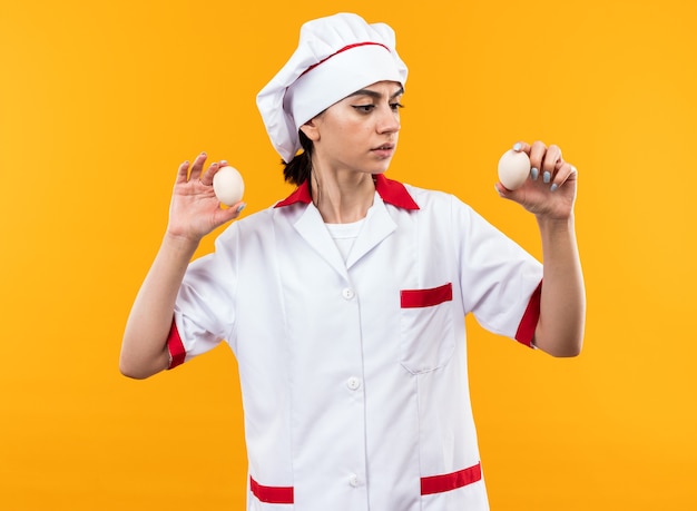 Pensando em uma jovem mulher bonita com uniforme de chef segurando e olhando para ovos isolados na parede laranja