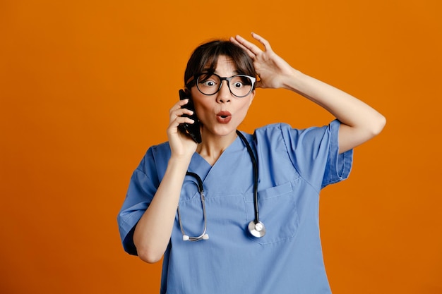 Pensando habla por teléfono joven doctora vistiendo uniforme fith estetoscopio aislado sobre fondo naranja