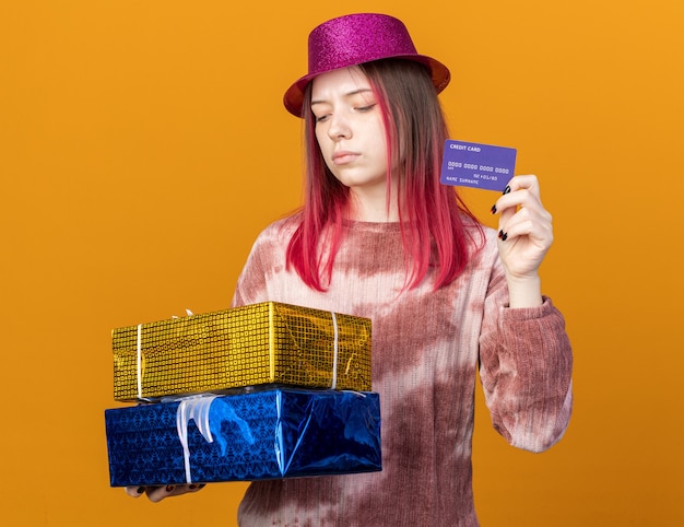 Pensando en la hermosa joven vistiendo gorro de fiesta sosteniendo cajas de regalo con tarjeta de crédito