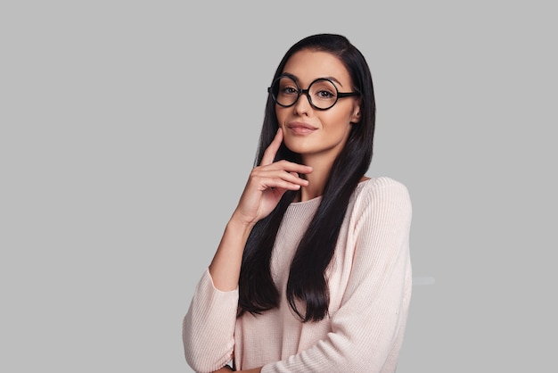 Pensando en ... una hermosa mujer joven sonriendo y mirando a la cámara mientras está de pie contra el fondo gris