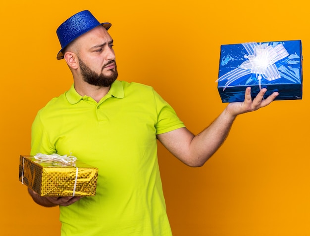 Pensando en el hombre joven con sombrero de fiesta sosteniendo y mirando cajas de regalo aisladas en la pared naranja