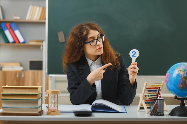 pensando jovem professora usando óculos segurando e aponta para fã número sentado na mesa com ferramentas escolares em sala de aula