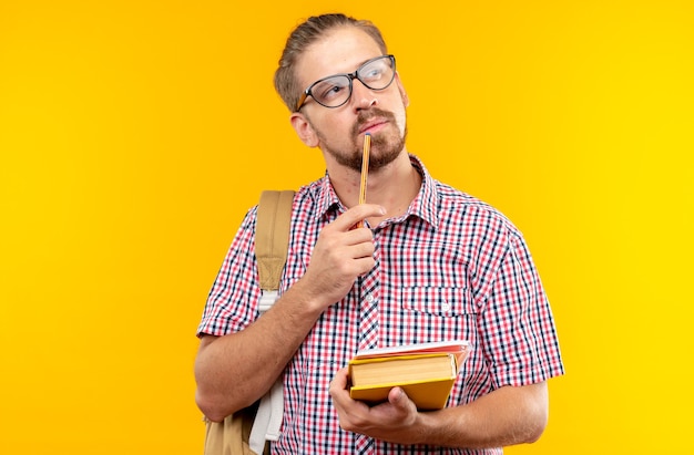 Pensando joven estudiante vistiendo mochila con gafas sosteniendo libro poniendo lápiz en la barbilla