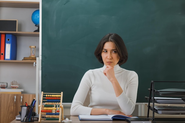 pensando mirando al lado agarró la barbilla joven maestra sentada en el escritorio con herramientas escolares en el aula