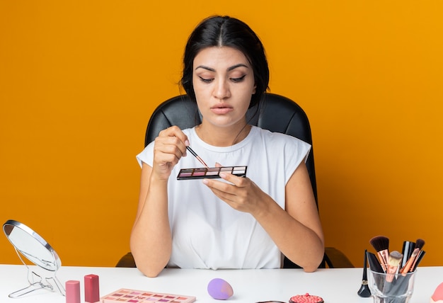 Pensando en una mujer hermosa se sienta a la mesa con herramientas de maquillaje sosteniendo y mirando la paleta de sombras de ojos con pincel de maquillaje