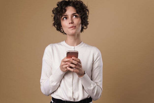 Pensando en una mujer hermosa usando un teléfono celular y mirando hacia arriba aislado sobre una pared beige