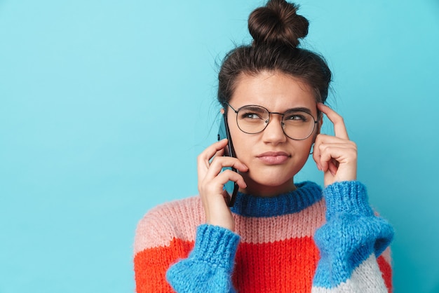 Pensando en la mujer joven en anteojos hablando por teléfono inteligente aislado sobre la pared azul