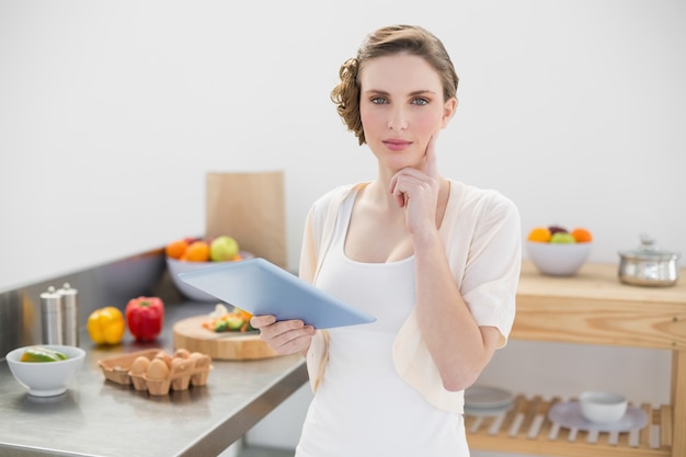 Foto pensando mulher deslumbrante de pé na cozinha enquanto segurava seu tablet