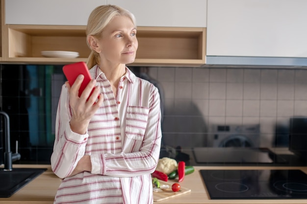 Pensando. Mulher em pé na cozinha com um smartphone na mão e parecendo pensativa