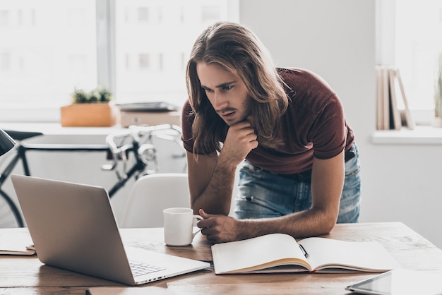 Pensando na solução. Jovem pensativo com cabelo comprido, olhando para seu laptop e segurando a mão no queixo enquanto se inclina para a mesa no escritório criativo