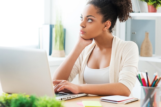 Pensando na solução. Mulher jovem e bonita africana trabalhando no laptop e olhando para longe enquanto está sentada em seu local de trabalho no escritório