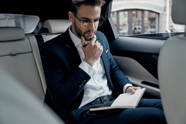 Foto pensando en negocios. apuesto joven en traje completo escribiendo algo en el organizador personal mientras está sentado dentro del coche