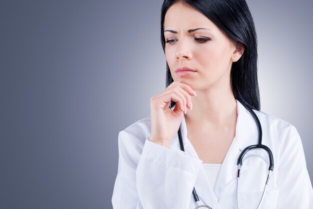 Pensando nesse problema. Médica de uniforme branco segurando a mão no queixo em pé contra um fundo cinza