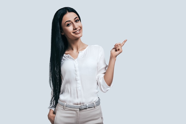 Pensando en nuevas soluciones. Mujer hermosa joven en camisa blanca apuntando hacia afuera y sonriendo mientras está de pie contra el fondo gris