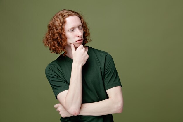 Pensando olhando para o lado agarrou o queixo jovem bonitão vestindo camiseta verde isolada em fundo verde