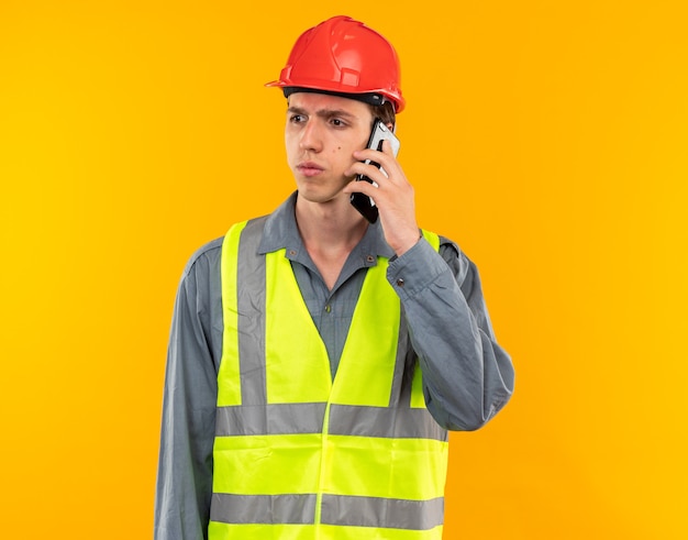 Pensando olhando para o lado, o jovem construtor de uniforme fala no telefone isolado na parede amarela