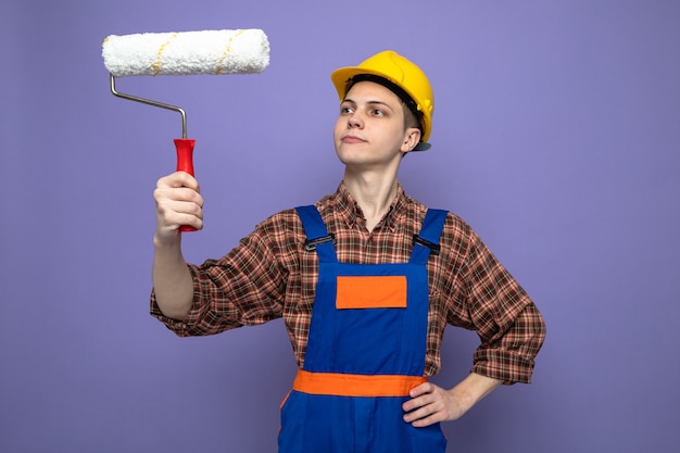 Pensando en poner la mano en la cadera joven constructor vistiendo uniforme sosteniendo y mirando el cepillo de rodillo