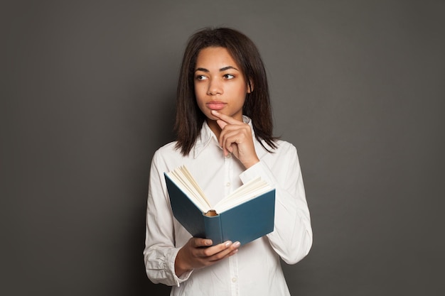 Pensando raza mixta etnia mujer negra estudiante en camisa blanca con libro en fondo gris