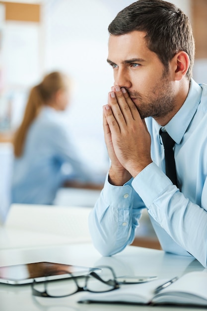 Pensando en la solución. Grave joven empresario en ropa formal manteniendo las manos juntas mientras está sentado en su lugar de trabajo