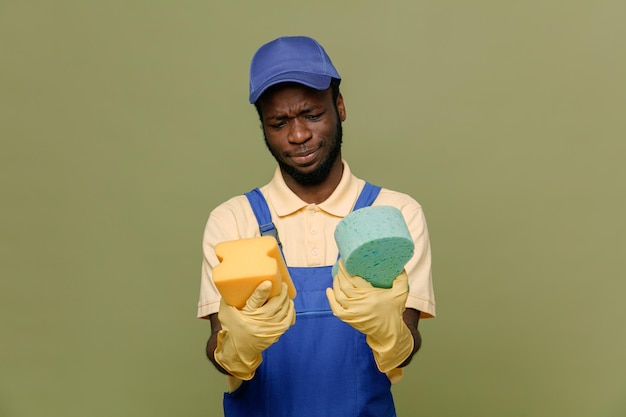 Pensando en sostener esponjas de limpieza joven limpiador afroamericano en uniforme con guantes aislados en fondo verde