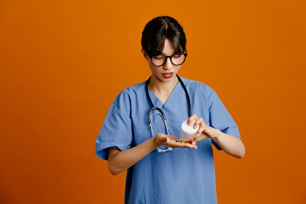 Pensando en sostener la píldora y el recipiente de la píldora joven doctora vistiendo uniforme fith estetoscopio aislado sobre fondo naranja