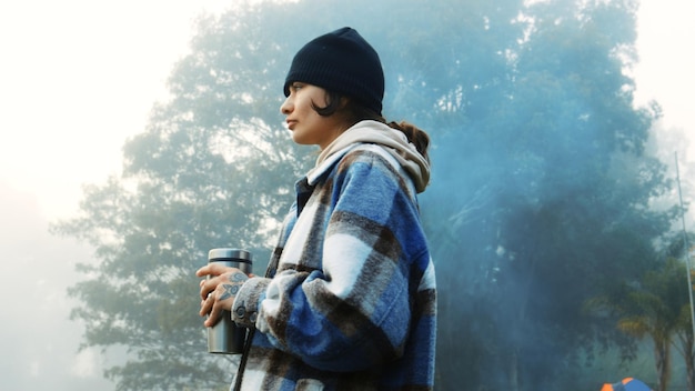 Pensar a natureza e uma mulher com café nas férias relaxam e acampam na floresta Ideia calma e uma jovem bebendo chá pela manhã em uma floresta durante um feriado no inverno para viagens