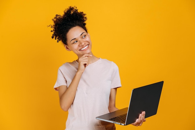 Foto pensativa afroamericana mujer de cabello rizado mira hacia otro lado pensativa piensa en una idea proyecto de puesta en marcha tiene una computadora portátil abierta en la mano se encuentra en un fondo naranja aislado sonríe soñando