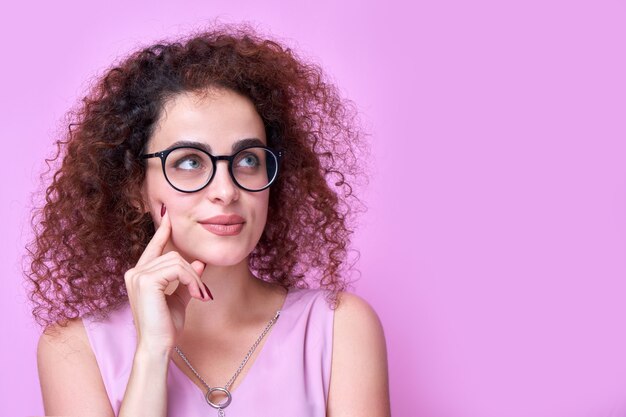 Pensativa chica armenia de pelo rizado con gafas piensa y toma decisiones, elige la solución al problema, mira a un lado perpleja, copia espacio sobre fondo rosa aislado