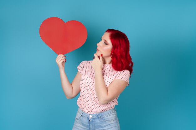 Foto pensativa e sonhadora jovem com cabelo vermelho segurando um grande coração de papel vermelho e olhando para ele