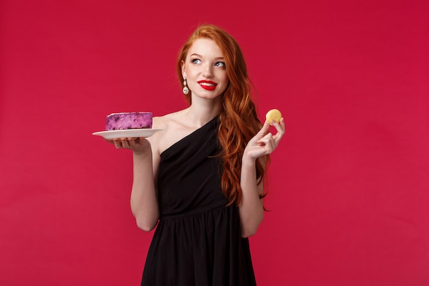 Pensativa e sonhadora linda ruiva magro de vestido preto, pensando em comer a sobremesa enquanto cuida de seu corpo, desviar o olhar ponderando e sorrindo, segure o biscoito e bolo